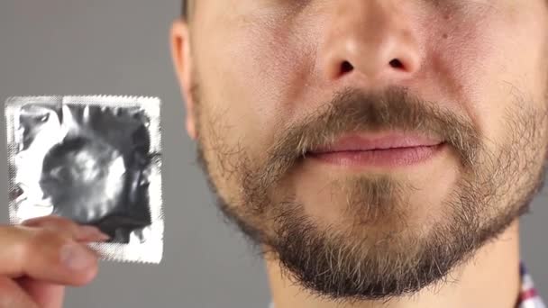 man met baard en snor houdt een nieuw condoom in de hand in de buurt van zijn gezicht en glimlacht, concept van een gezonde levensstijl en seksuele relaties, grijze achtergrond, vooraanzicht  - Video