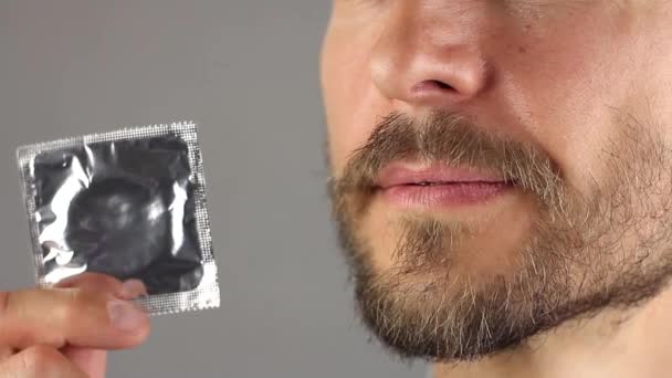 man met baard en snor houdt nieuw condoom in de hand in de omgeving van die zijn gezicht verheugt zich en glimlacht, zijaanzicht, concept van een gezonde levensstijl en seksuele relaties, grijze achtergrond  - Video