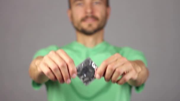 man in green shirt raises in front of him on his outstretched hands new condom in square package, holding it by corners, portrait of man is blurred - not in focus, concept of healthy lifestyle, gray background   - Footage, Video