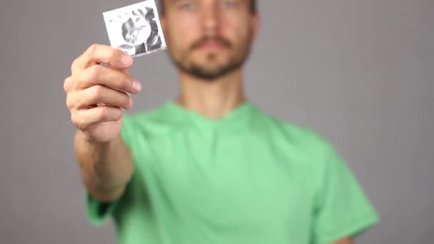 Mann im grünen Hemd hebt vor sich auf der rechten ausgestreckten Hand neues Kondom, linke Hand macht Geste Daumen hoch, Freude und Vorfreude auf Vergnügen, angemessene Haltung zum Schutz, grauer Hintergrund  - Filmmaterial, Video