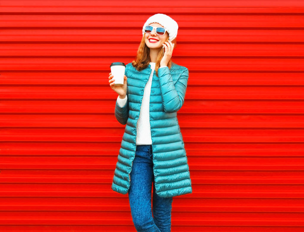 Fashion smiling woman talks on a smartphone holds coffee cup on a red background in the city - Photo, Image