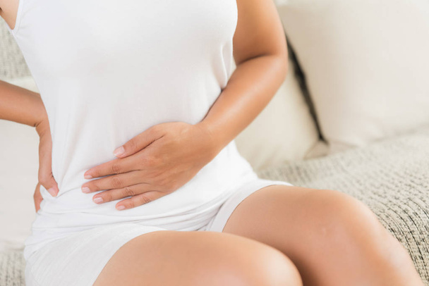 Mujer joven con dolor de estómago y dolor de espalda sentada en el sofá en casa. Gastritis crónica. Concepto de distensión abdominal
. - Foto, Imagen