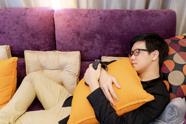 Азіатський юнак спати на Сафа обіймати подушка утримуючи пульт дистанційного керування ТВ в кімнаті домашнього кінотеатру. Головна живуть способу життя концепція - Фото, зображення