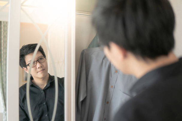 Jeune homme asiatique regardant dans le miroir sur le placard tout en choisissant une chemise de style décontracté pour s'habiller dans la chambre. Concept de vie à domicile
 - Photo, image