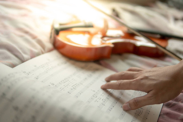 Musician hand on sheet of music note and vintage violin on flowery pink bed in the bedroom. Playing music instrument at home concept - Photo, Image