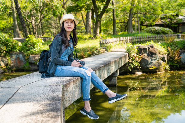 Młoda atrakcyjna kobieta sittign na moście w pięknym ogrodzie z pełnym roślin i rzeki. - Zdjęcie, obraz