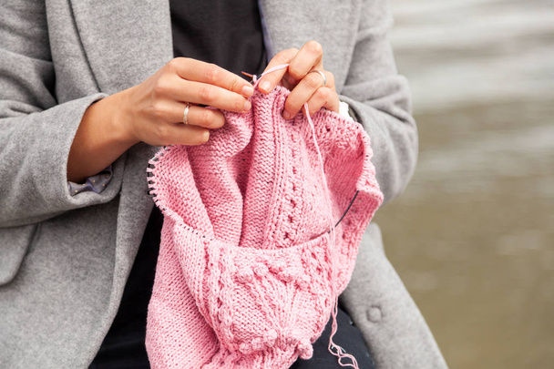 Close up og a young  married woman dressed in trendy  coat knitting a pink hat with knitting needles  against  sea  in sunny autumn day . Freelance creative working and living concept - Foto, Imagen