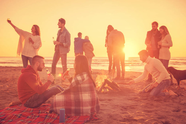 Mladý pár těší s přáteli kolem táboráku na pláži při západu slunce pití piva - Fotografie, Obrázek