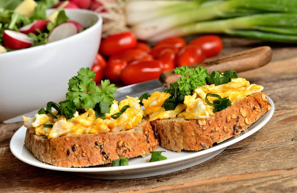 Close-up of wholemeal bread with scrambled eggs, fresh herbs, spring onions, tomatoes and salad bowl in background - healthy breakfast - Photo, Image