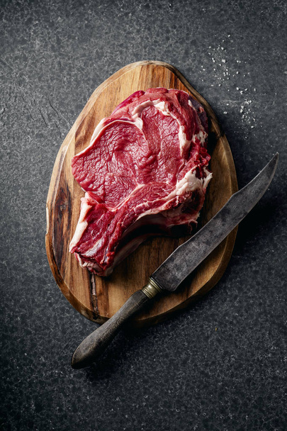 Top view of raw beef steak on wooden chopping board with knife on black concrete background - Фото, зображення