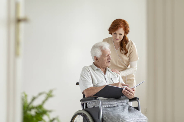 Caregiver supporting paralyzed elderly man in a wheelchair with photo album - Zdjęcie, obraz