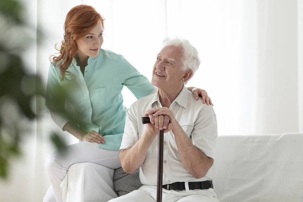 Friendly nurse and smiling elderly man with walking stick in the nursing house - Photo, image
