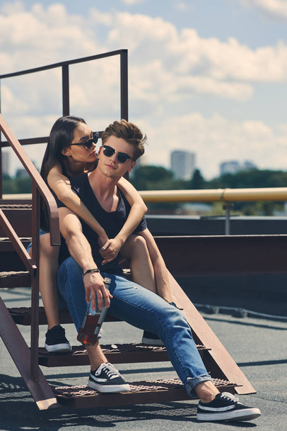 young multiethnic couple with bottle of whiskey sitting on stairs on roof - Foto, immagini