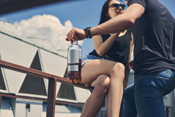 hot multicultural couple flirting and holding bottle of rum on roof - Photo, Image