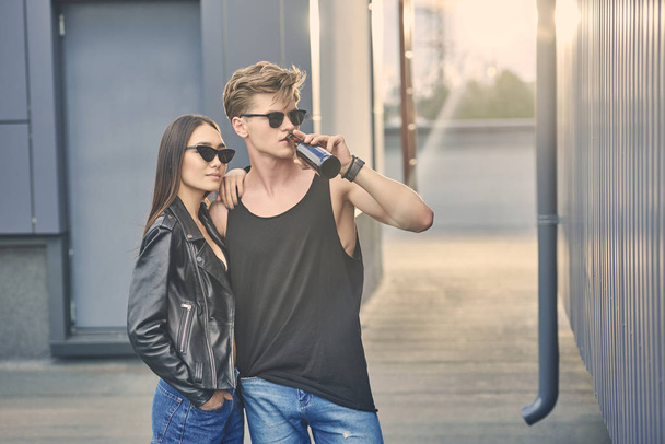 multikulturní pár v trendy brýle objímání na střeše s podsvícením, člověk pít pivo - Fotografie, Obrázek