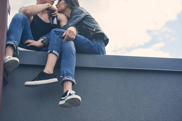 stylish multiethnic couple with bottle of beer embracing and sitting on roof  - Photo, Image