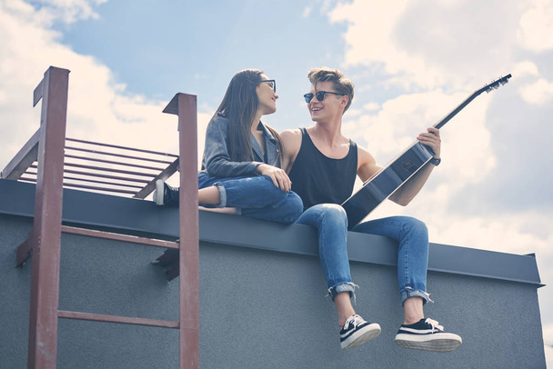 happy multicultural couple sitting on roof with acoustic guitar - Foto, afbeelding
