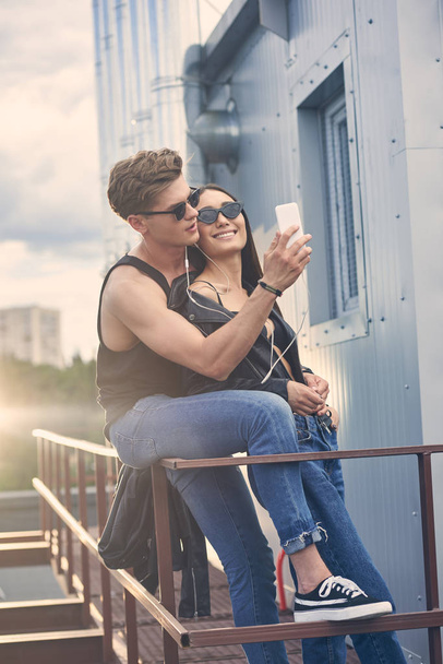 happy interracial couple listening music with earphones and taking selfie on smartphone on roof - Foto, Imagem