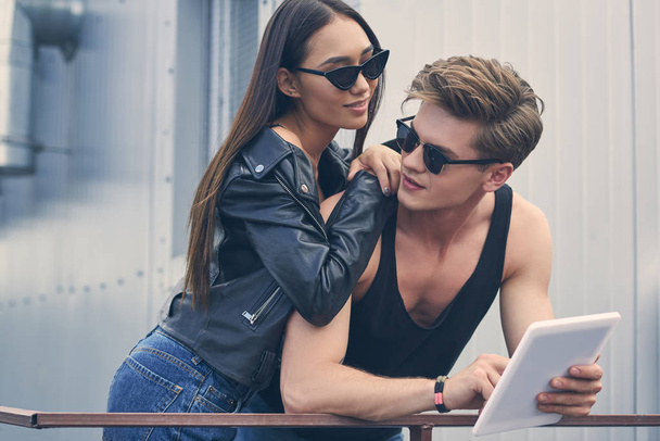 молода багатоетнічна пара в сонцезахисних окулярах обіймається і використовує цифровий планшет
  - Фото, зображення