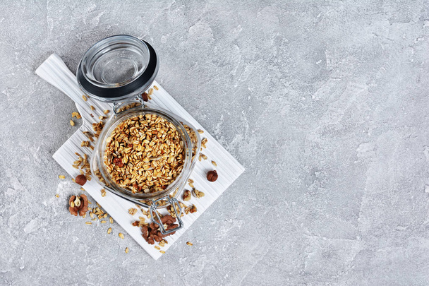 Top view on healthy breakfast homemade granola with nuts and sunflower seeds in open glass jar on white wooden board and gray concrete background - Φωτογραφία, εικόνα
