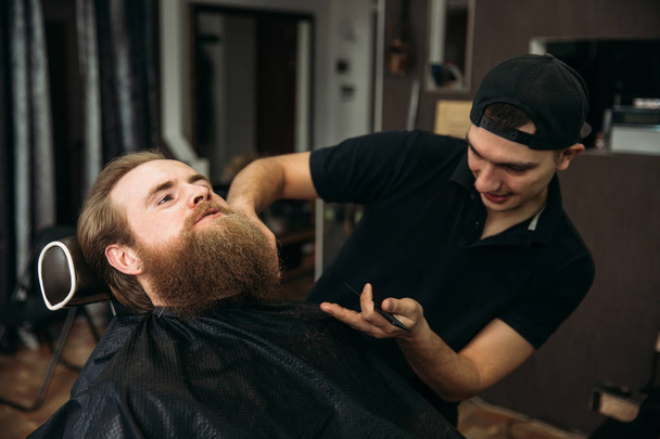 The hands of young barber making haircut to attractive bearded man in barbershop - Photo, Image