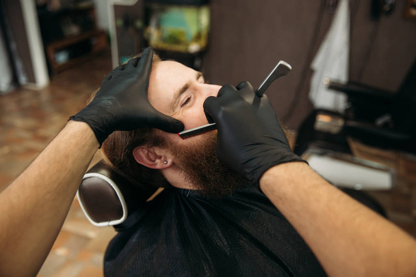 Бородатий чоловік з довгою бородою отримує стильне гоління волосся, стрижка, з бритвою перукаря в перукарні
 - Фото, зображення