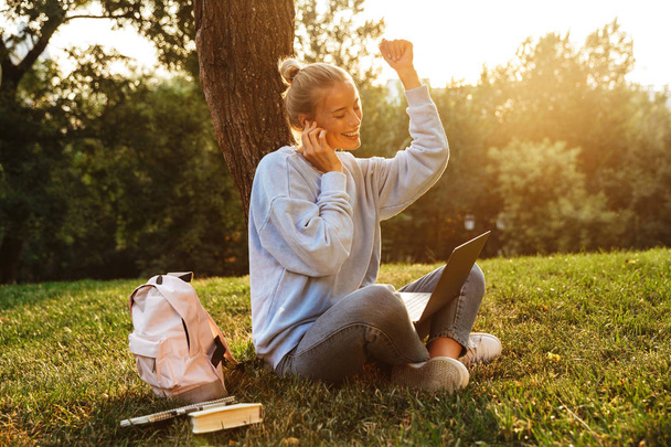 Portrait of a cheerful young girl in earphones with backpack sitting with legs crossed on a grass at the park, using laptop computer - Fotografie, Obrázek