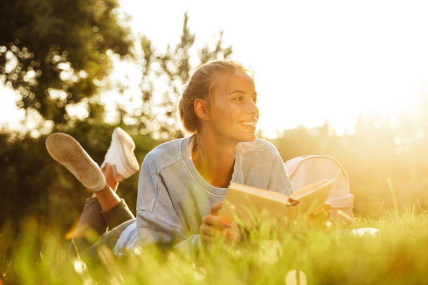Portrét šťastné mladé dívky s batohem na trávě v parku, čtení knihy - Fotografie, Obrázek