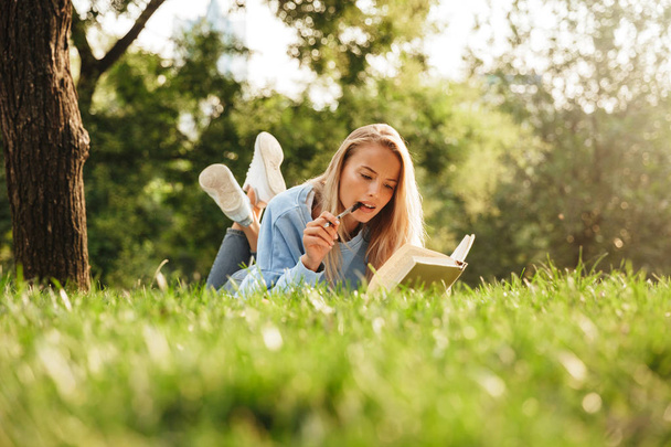 Portrait of a pensive young girl laying on a grass at the park, reading a book, taking notes - Foto, imagen
