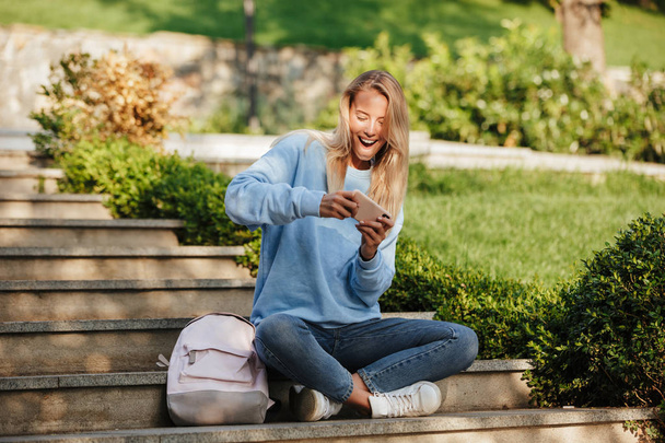 Portrait of a happy young girl student with backpack sitting on steps outdoors, playing games on mobile phone - Φωτογραφία, εικόνα