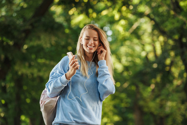 Étudiante souriante avec sac à dos tenant un téléphone portable, marchant dans le parc, écoutant de la musique avec des écouteurs
 - Photo, image