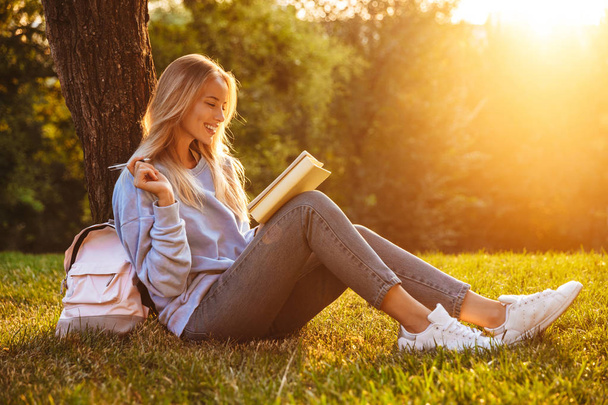 Portrait of a happy young girl sitting on a grass at the park, reading a book, taking notes - Foto, Imagem
