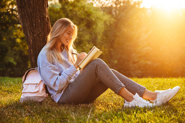 Portrait of a cheerful young girl sitting on a grass at the park, reading a book, taking notes - Photo, Image