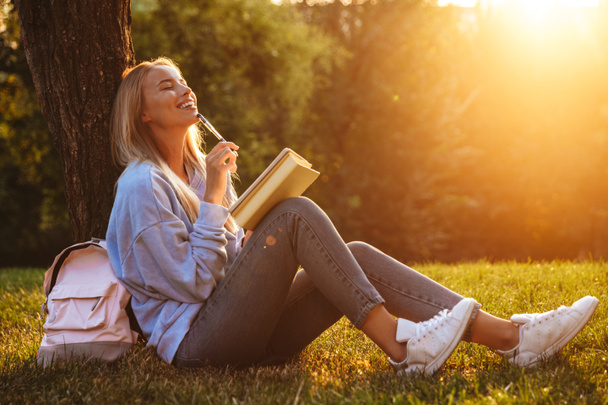 Πορτραίτο από ένα ονειρικό νεαρό κορίτσι που κάθεται σε ένα χόρτο στο πάρκο, διαβάζοντας ένα βιβλίο, να κρατάς σημειώσεις - Φωτογραφία, εικόνα