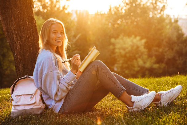 Portrait of a happy young girl sitting on a grass at the park, reading a book, taking notes - Foto, imagen