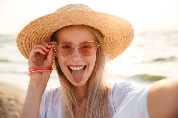 Cheerful young girl in summer hat and swimwear spending time at the beach, taking a selfie - Foto, imagen