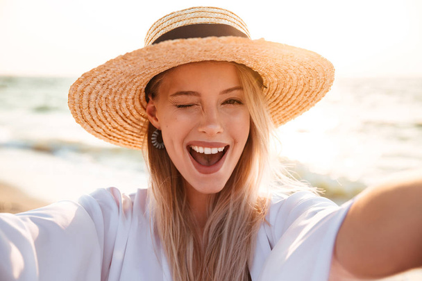 Portrait of lovely pleased blonde woman 20s in summer straw hat smiling and taking selfie while walking at sea coast - Foto, afbeelding