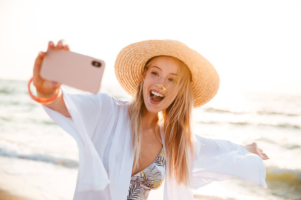 Excited young girl in summer hat and swimwear spending time at the beach, taking a selfie with outsretched hand - Foto, Imagen