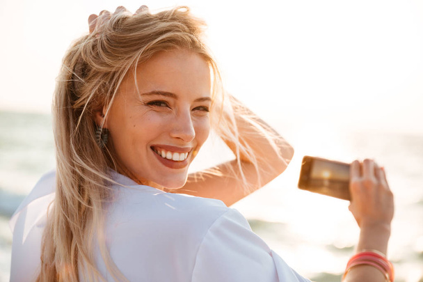 Photo of joyous blonde woman 20s smiling, and taking photo of sunrise above sea while walking on summer beach - Photo, Image