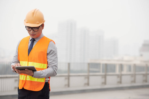 Young Asian construction supervisor reading information in tablet computer - Photo, Image