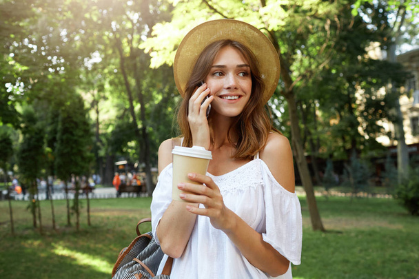 Открытый портрет счастливой милой молодой женщины носит стильную летнюю шляпу, рюкзак и белое платье, разговаривает по мобильному телефону и пьет кофе на вынос в парке в городе
 - Фото, изображение