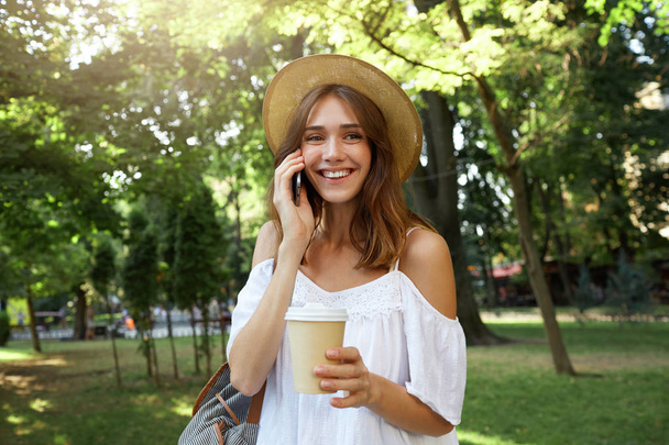Outdoor portrait of cheerful lovely young woman wears stylish summer hat and white dress, feels relaxed, talking on cell phone and drinking takeaway coffee in park - Photo, Image