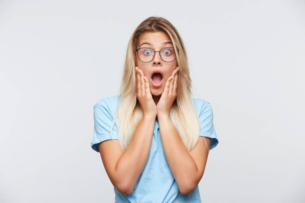 Portrait of shocked stunned young woman with blonde hair and opened mouth wears glasses and blue t shirt keeps hands on cheeks and feels amazed isolated over white background - Photo, Image