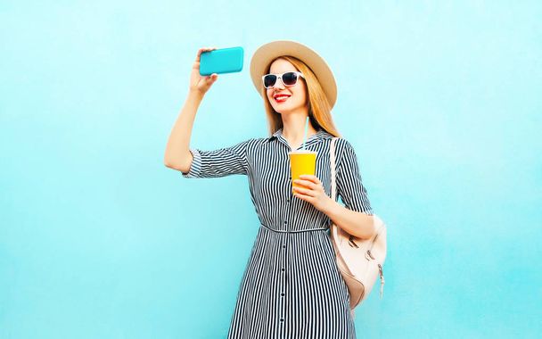 Fashion smiling woman takes a picture self portrait on smartphone holds cup of juice with backpack on blue background - Foto, Imagem