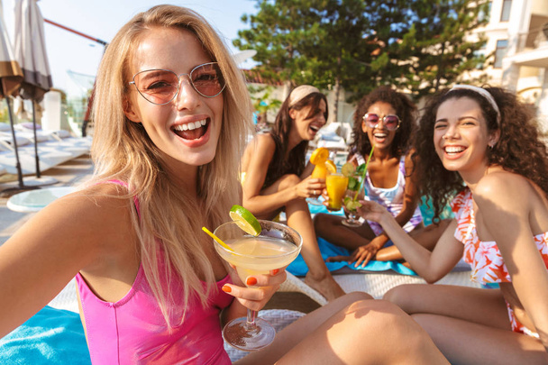 Image of young happy emotional group of women friends drinking cocktails sitting outdoors in pool dressed in swimwears take a selfie by camera. - Фото, зображення