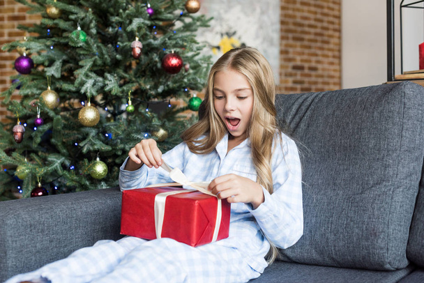 surprised child in pajamas opening christmas gift while sitting on couch - Fotoğraf, Görsel