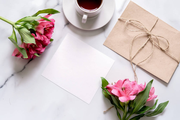 Marble desk with cup of coffee, pink flowers, postcard, kraft envelope, twine, cotton branch, oat cookies, invitation card with copy space for text. Flat lay, top view. Woman's day or mother's day. - Φωτογραφία, εικόνα