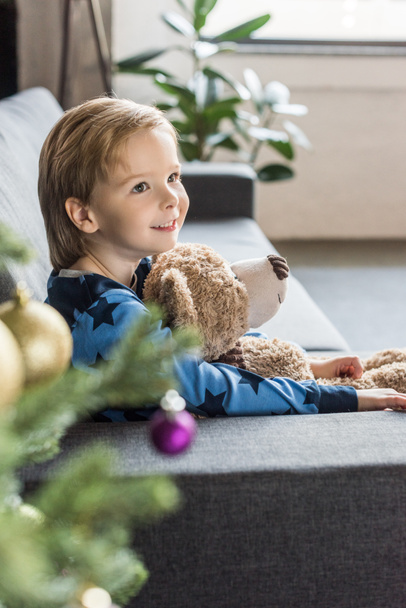 vista lateral de lindo niño sonriente sosteniendo oso de peluche y sofá sentado
 - Foto, Imagen