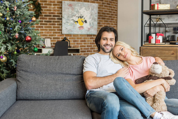 schönes glückliches junges Paar sitzt zusammen auf dem Sofa und lächelt an Heiligabend in die Kamera - Foto, Bild