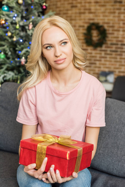beautiful young blonde woman holding christmas present and looking away at home - Zdjęcie, obraz
